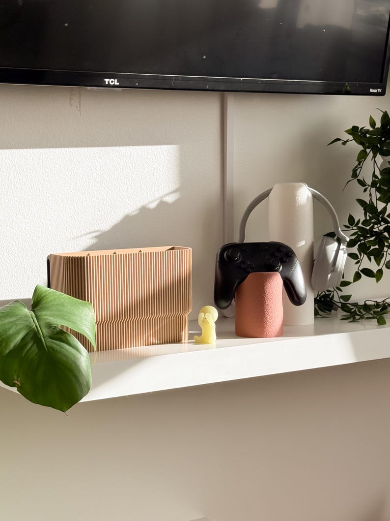 May include: A wooden rectangular box with vertical lines, a small yellow figurine, a black game controller, and a pink cylindrical object are on a white shelf. A pair of gray headphones and a green plant are also on the shelf. A black television is in the background.