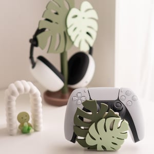 May include: A white and gray game controller with a green monstera leaf design. A matching headset stand with a monstera leaf design is behind the controller. A small white arch with a green figure is in the foreground.