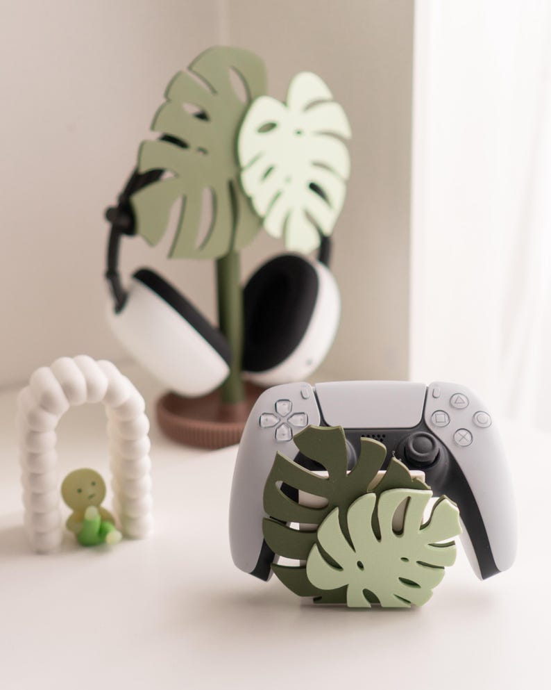 May include: A white and gray game controller with a green monstera leaf design. A matching headset stand with a monstera leaf design is behind the controller. A small white arch with a green figure is in the foreground.
