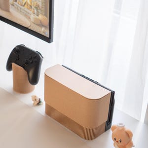May include: A light brown console with a matching controller stand and a black controller. A small plush bear and a figurine are also present. The console has a curved top and a ribbed texture on the front. The controller stand has a ribbed design.
