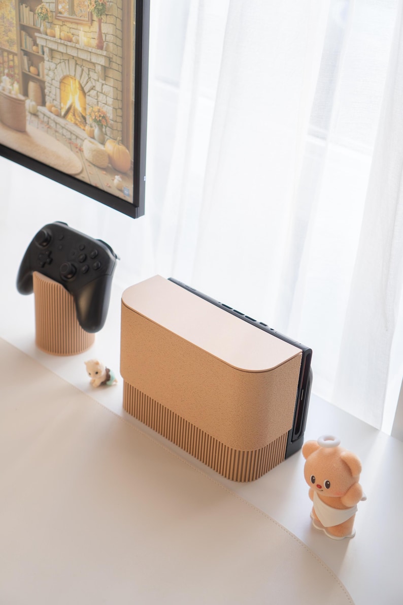 May include: A light brown console with a matching controller stand and a black controller. A small plush bear and a figurine are also present. The console has a curved top and a ribbed texture on the front. The controller stand has a ribbed design.
