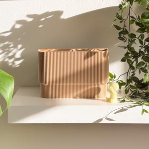 May include: A light brown, rectangular storage container with vertical ribbed detailing. The container sits on a white shelf, with a small yellow figurine and trailing green plant in the background. The container is likely for home decor.