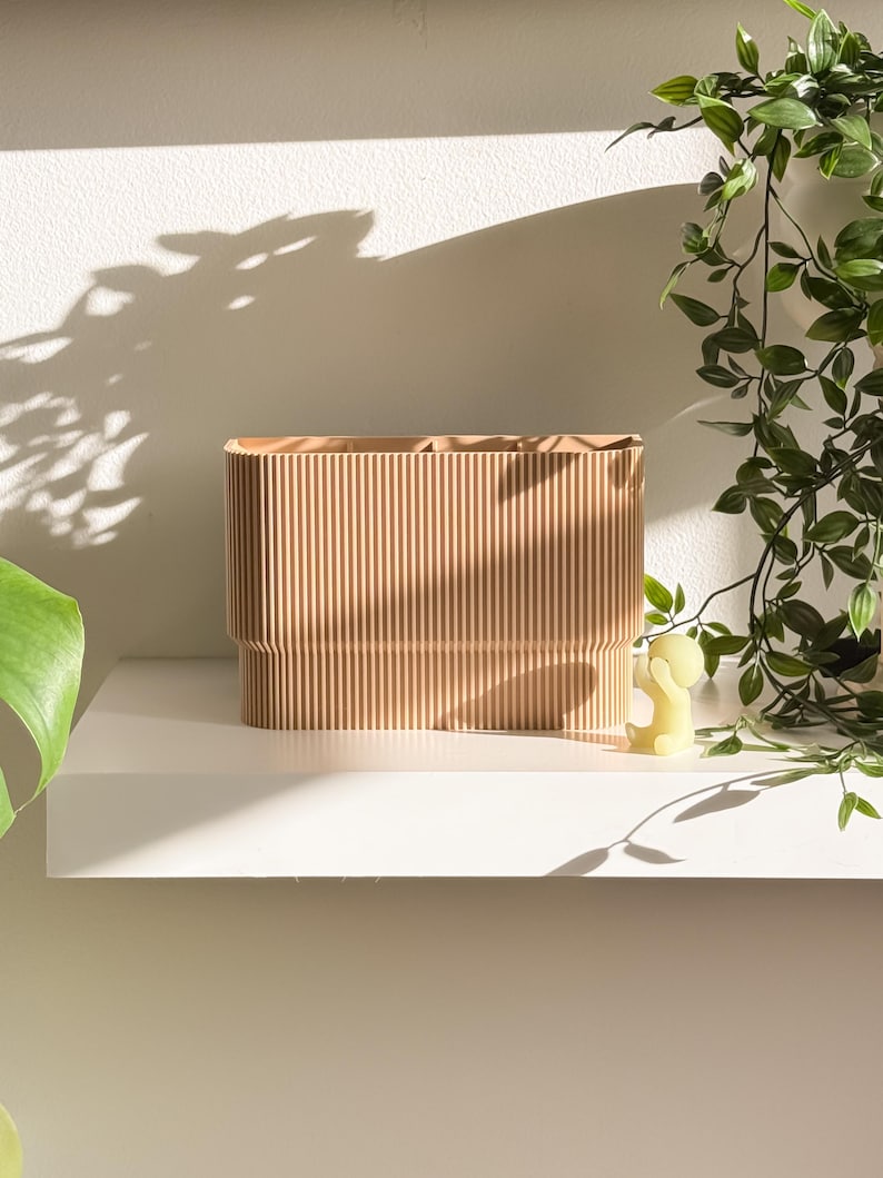 May include: A light brown, rectangular storage container with vertical ribbed detailing. The container sits on a white shelf, with a small yellow figurine and trailing green plant in the background. The container is likely for home decor.