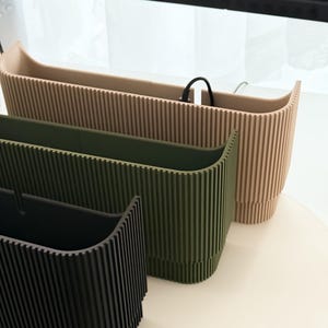 May include: Three modern, ribbed planters in a row. The planters are in shades of black, olive green, and beige. The planters have a rectangular shape with a curved top edge. The planters are made of a solid material and are suitable for indoor or outdoor use.