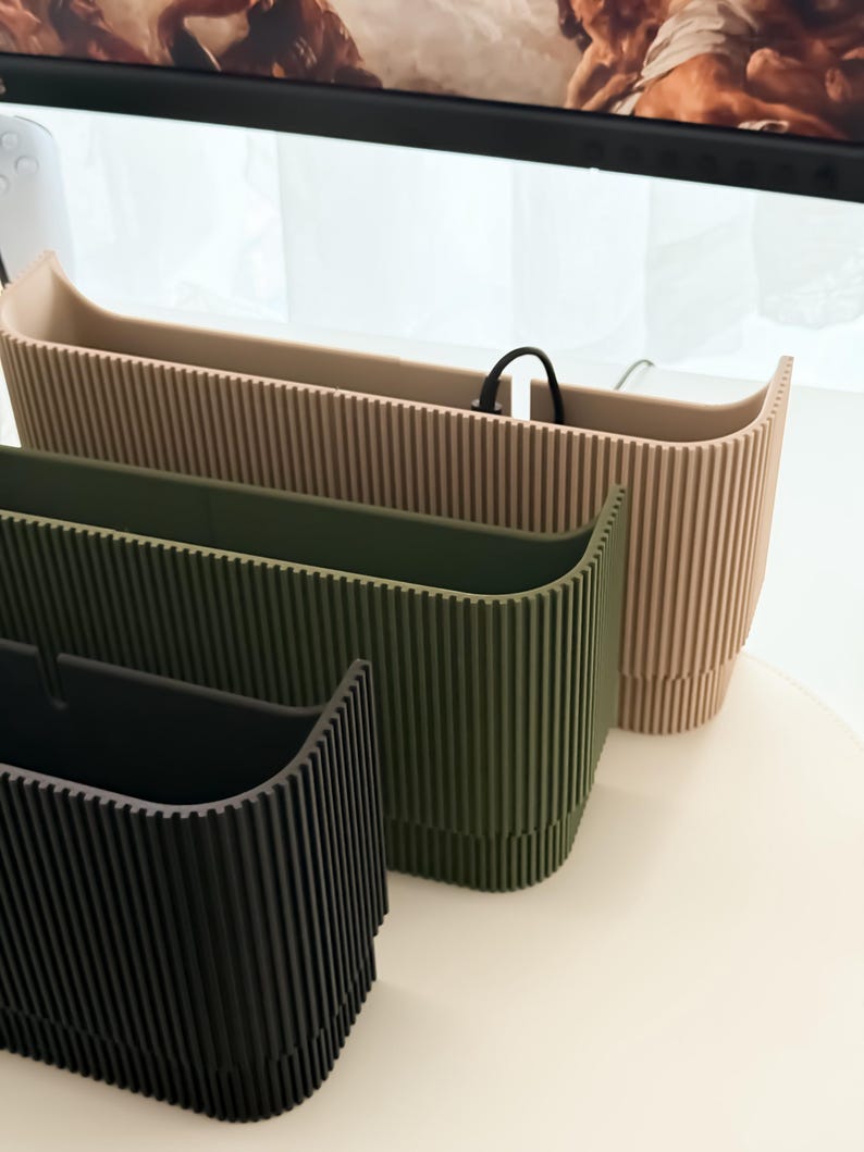 May include: Three modern, ribbed planters in a row. The planters are in shades of black, olive green, and beige. The planters have a rectangular shape with a curved top edge. The planters are made of a solid material and are suitable for indoor or outdoor use.