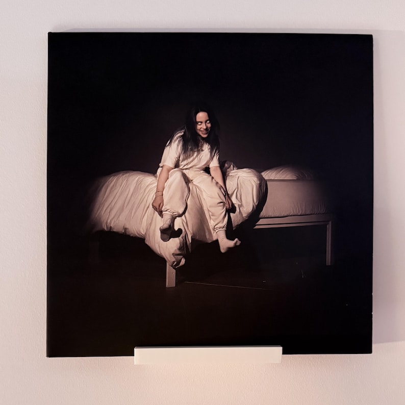 May include: A framed album cover featuring a person sitting on a bed. The person is wearing a white outfit and smiling. The background is dark, creating a spotlight effect on the bed and the person.
