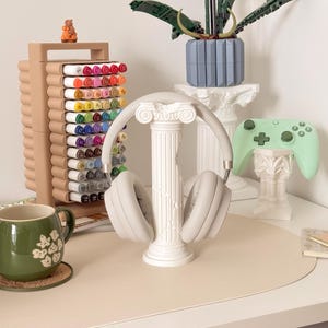Puede incluir: Una configuraci&oacute;n de escritorio con unos auriculares gris claro sobre un soporte de columna blanco. Tambi&eacute;n se ven una taza verde, un organizador de rotuladores con rotuladores de colores y un mando de juego verde menta. Una orqu&iacute;dea Lego y una peque&ntilde;a figura completan la escena.