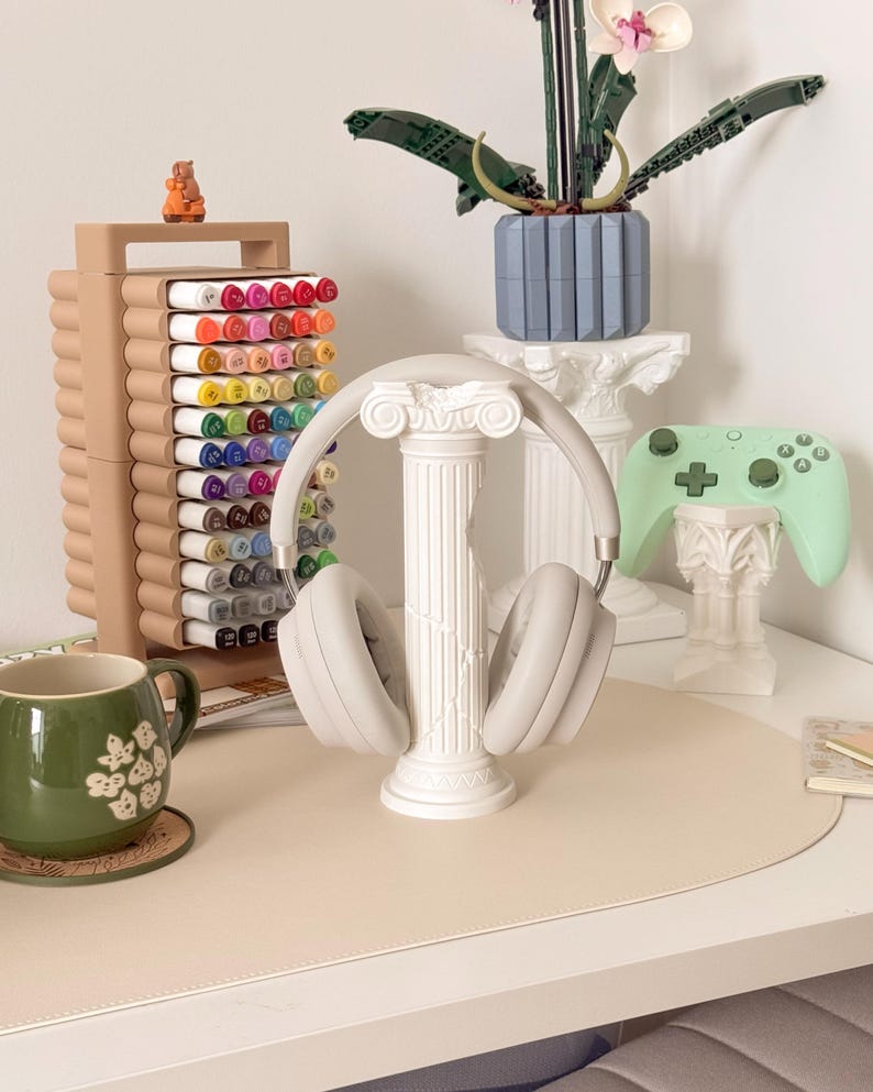 Puede incluir: Una configuraci&oacute;n de escritorio con unos auriculares gris claro sobre un soporte de columna blanco. Tambi&eacute;n se ven una taza verde, un organizador de rotuladores con rotuladores de colores y un mando de juego verde menta. Una orqu&iacute;dea Lego y una peque&ntilde;a figura completan la escena.