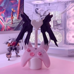 May include: A pink and black gaming controller stand with a heart-shaped top and two wings. The stand is holding a pink Xbox controller.