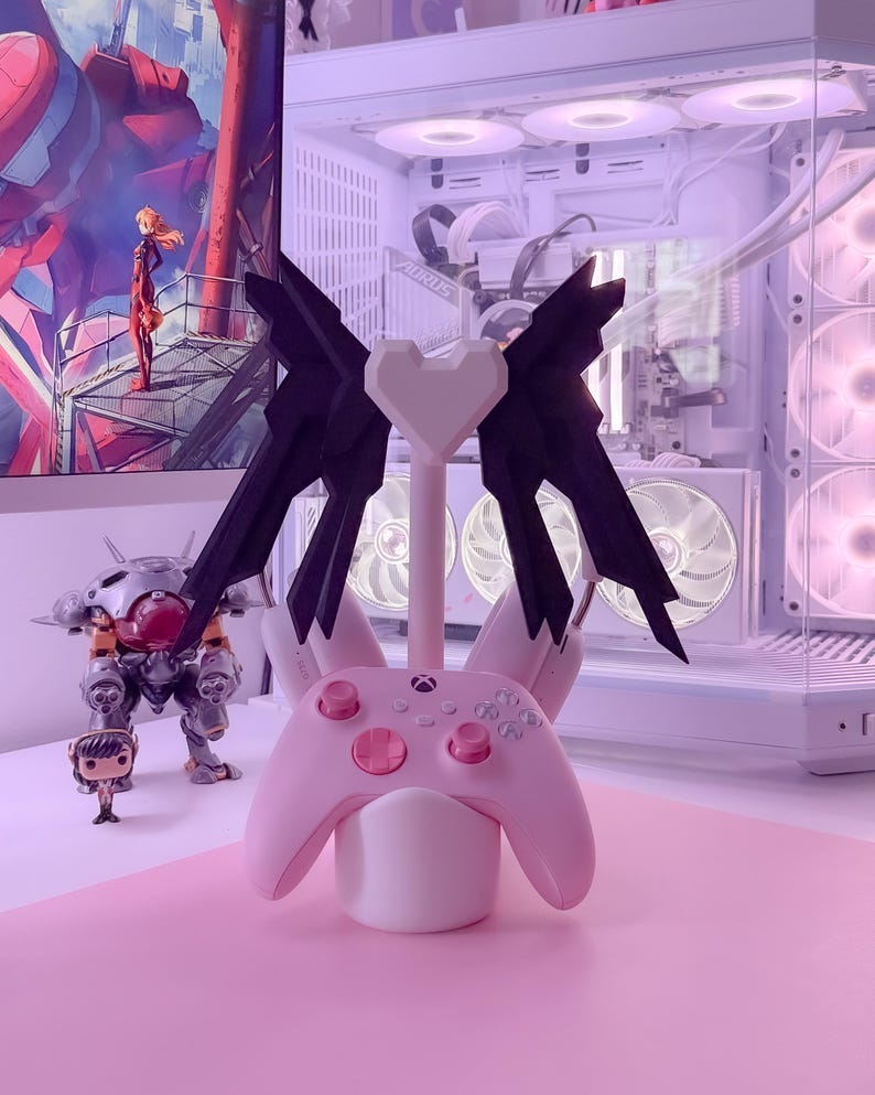 May include: A pink and black gaming controller stand with a heart-shaped top and two wings. The stand is holding a pink Xbox controller.