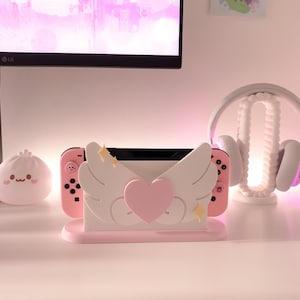 May include: A pink and white Nintendo Switch console stand with a heart and wings design. The stand has a pink heart in the center and two white wings on either side. There are two gold stars on each wing. The stand is on a white surface.