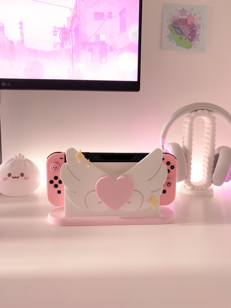 May include: A pink and white Nintendo Switch console stand with a heart and wings design. The stand has a pink heart in the center and two white wings on either side. There are two gold stars on each wing. The stand is on a white surface.