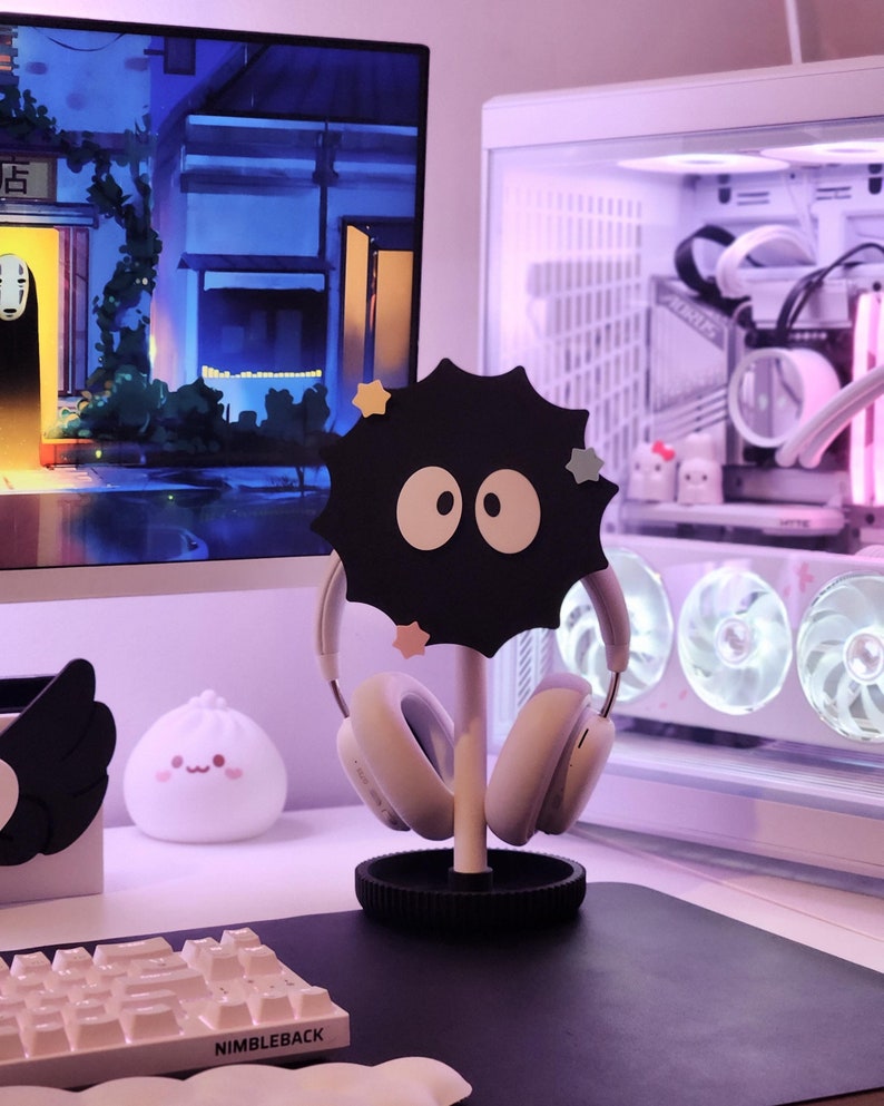 May include: A black headphone stand featuring a cartoon soot sprite design, supporting white headphones. The stand has a circular base and a central pole. The background includes a computer monitor, a computer case, and a keyboard.
