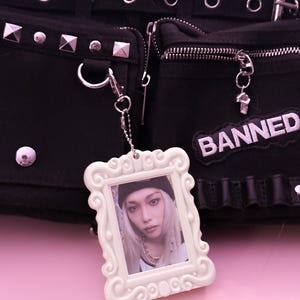 May include: Black bag with silver hardware, studs, and zippers. A white framed photo charm hangs from the bag. The charm features a person with long blonde hair. A patch that says "BANNED" is on the bag.