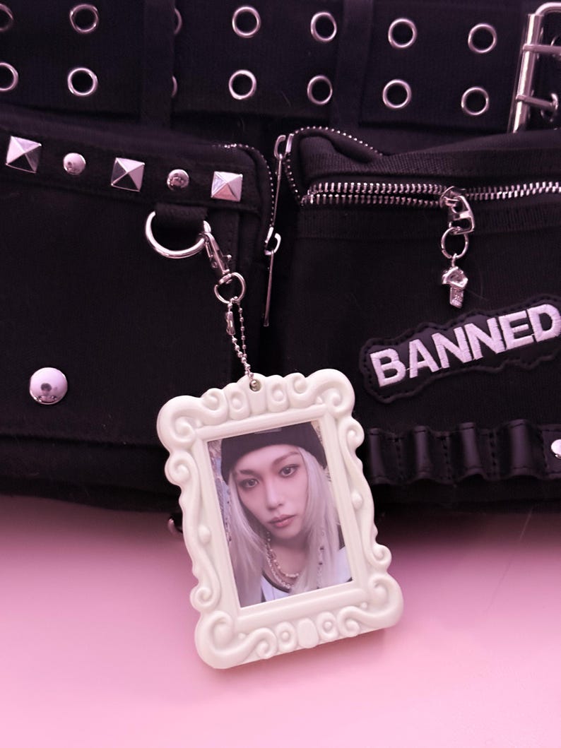 May include: Black bag with silver hardware, studs, and zippers. A white framed photo charm hangs from the bag. The charm features a person with long blonde hair. A patch that says "BANNED" is on the bag.