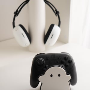 May include: A black and white Nintendo Switch Pro Controller with a white ghost-shaped stand. In the background, a pair of white and black headphones rest on a white cylindrical stand. The items are on a white surface.