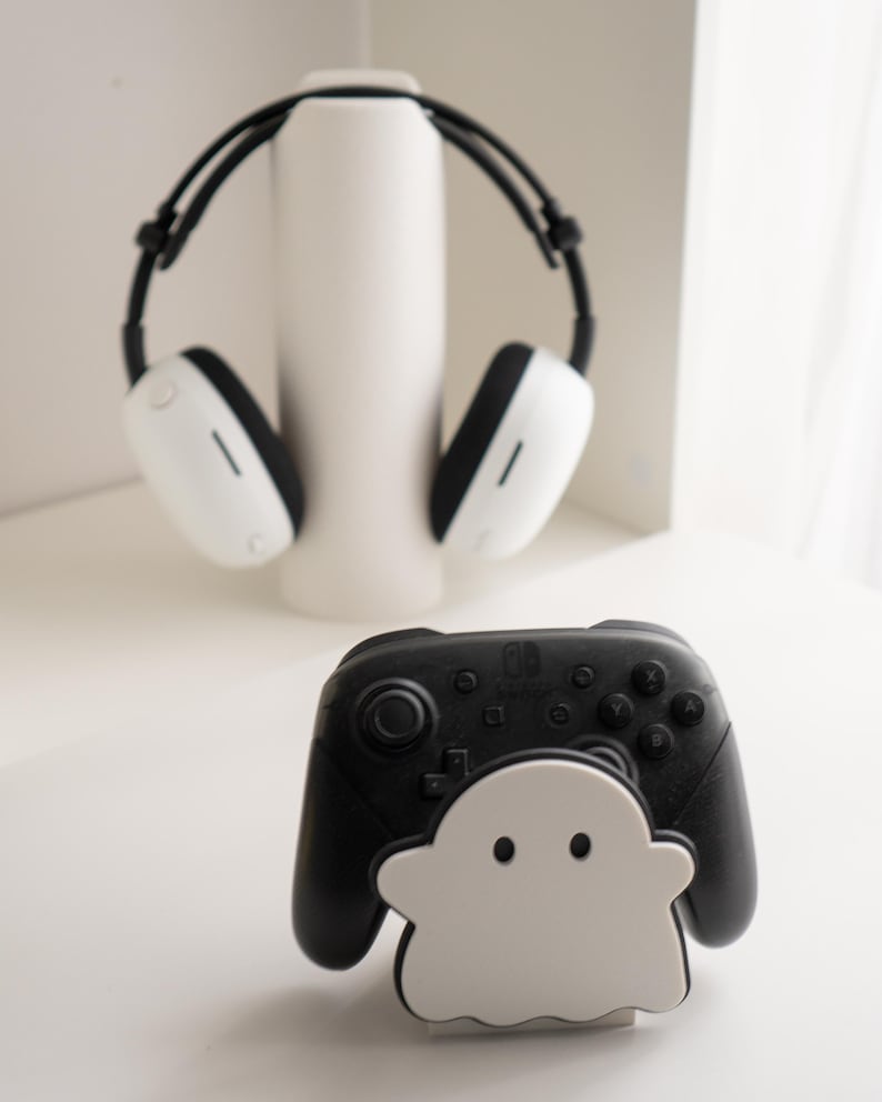 May include: A black and white Nintendo Switch Pro Controller with a white ghost-shaped stand. In the background, a pair of white and black headphones rest on a white cylindrical stand. The items are on a white surface.