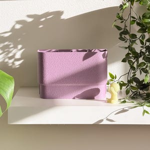 May include: A textured, rectangular lavender planter sits on a white shelf. The planter has a layered design and is empty. A small yellow candle shaped like a person is next to the planter. Green leafy vines hang from above.
