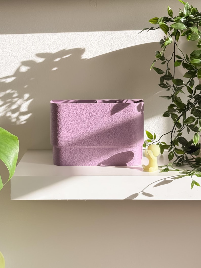 May include: A textured, rectangular lavender planter sits on a white shelf. The planter has a layered design and is empty. A small yellow candle shaped like a person is next to the planter. Green leafy vines hang from above.