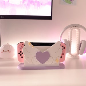 May include: A pink and white Nintendo Switch console stand with a purple heart and white wings. The stand has a light purple base.