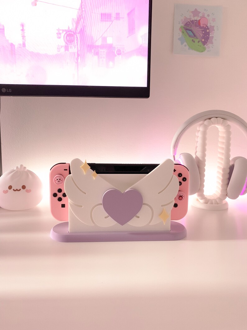 May include: A pink and white Nintendo Switch console stand with a purple heart and white wings. The stand has a light purple base.