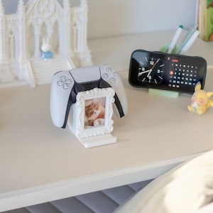 May include: A white plastic controller stand featuring an ornate, rectangular picture frame design. The stand holds a white and black gaming controller and displays a small photograph of a brown and white cat inside the frame.