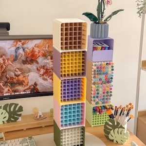 May include: A vertical stack of five square modular desk organizers in cream, purple, yellow, pink, and olive green, each featuring a grid interior. Beside it, a second stack holds markers and pens in light purple, beige, and light green containers.