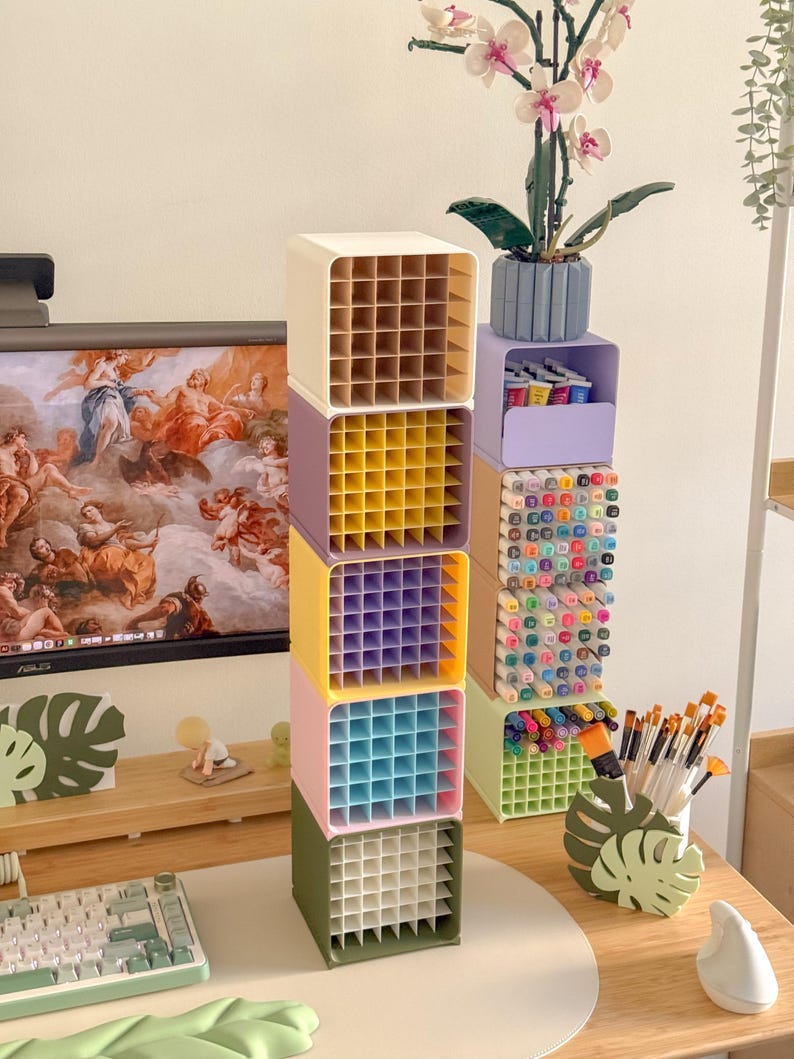 May include: A vertical stack of five square modular desk organizers in cream, purple, yellow, pink, and olive green, each featuring a grid interior. Beside it, a second stack holds markers and pens in light purple, beige, and light green containers.