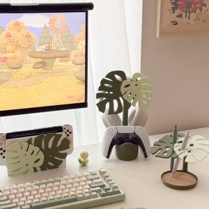 Pu&ograve; includere: Un'impostazione di gioco con una tastiera bianca, una Nintendo Switch e un controller PlayStation 5, tutti decorati con disegni di foglie di monstera verdi. Sono presenti anche una piccola statuina gialla e un portapenne a forma di foglia di monstera.