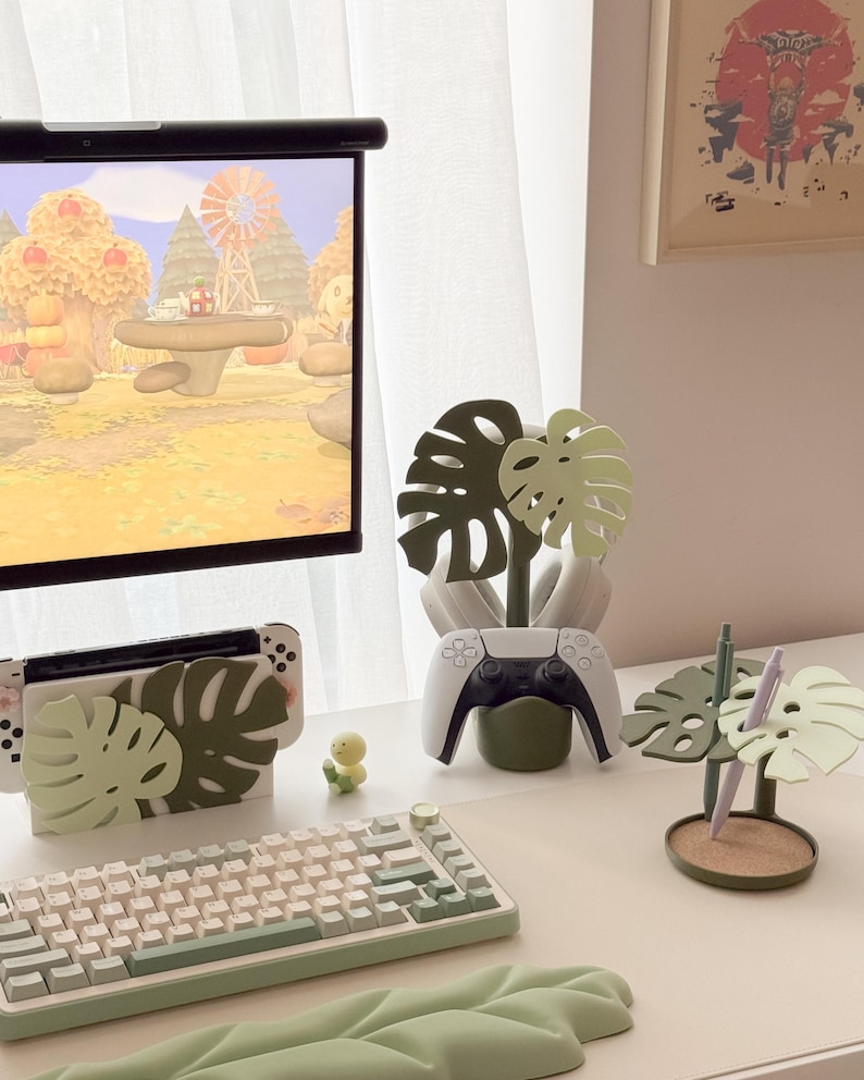 Pu&ograve; includere: Un'impostazione di gioco con una tastiera bianca, una Nintendo Switch e un controller PlayStation 5, tutti decorati con disegni di foglie di monstera verdi. Sono presenti anche una piccola statuina gialla e un portapenne a forma di foglia di monstera.