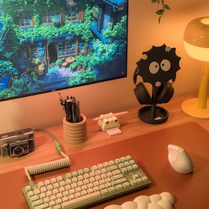 May include: A contemporary desk setup with a computer monitor showing a scenic image, a green and white keyboard, a white mouse, and a black headphone stand. Other items include a vintage camera, a pencil holder, and a yellow lamp.