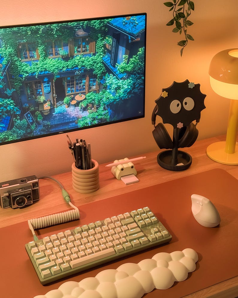 May include: A contemporary desk setup with a computer monitor showing a scenic image, a green and white keyboard, a white mouse, and a black headphone stand. Other items include a vintage camera, a pencil holder, and a yellow lamp.