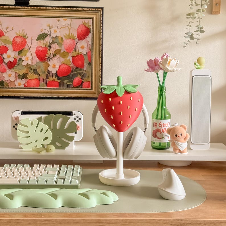 May include: A red strawberry-shaped headphone stand with a green stem and leaves, holding white headphones. The stand is on a white base, with a keyboard, mouse, and other decorative items in the background. A painting of strawberries hangs on the wall.