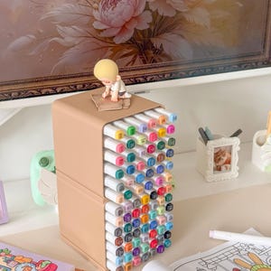 May include: A beige plastic storage organizer holding a large collection of multi-colored markers arranged in a grid. A small blonde figurine sits on top of the unit. The markers feature white caps with color-coded ends and printed numbers.