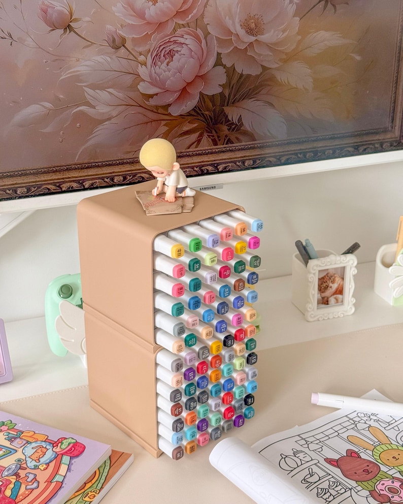 May include: A beige plastic storage organizer holding a large collection of multi-colored markers arranged in a grid. A small blonde figurine sits on top of the unit. The markers feature white caps with color-coded ends and printed numbers.