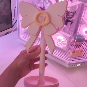 May include: A pink and white decorative stand with a large bow and a small yellow circle in the center. The stand has a round base and a tall, slender stem.