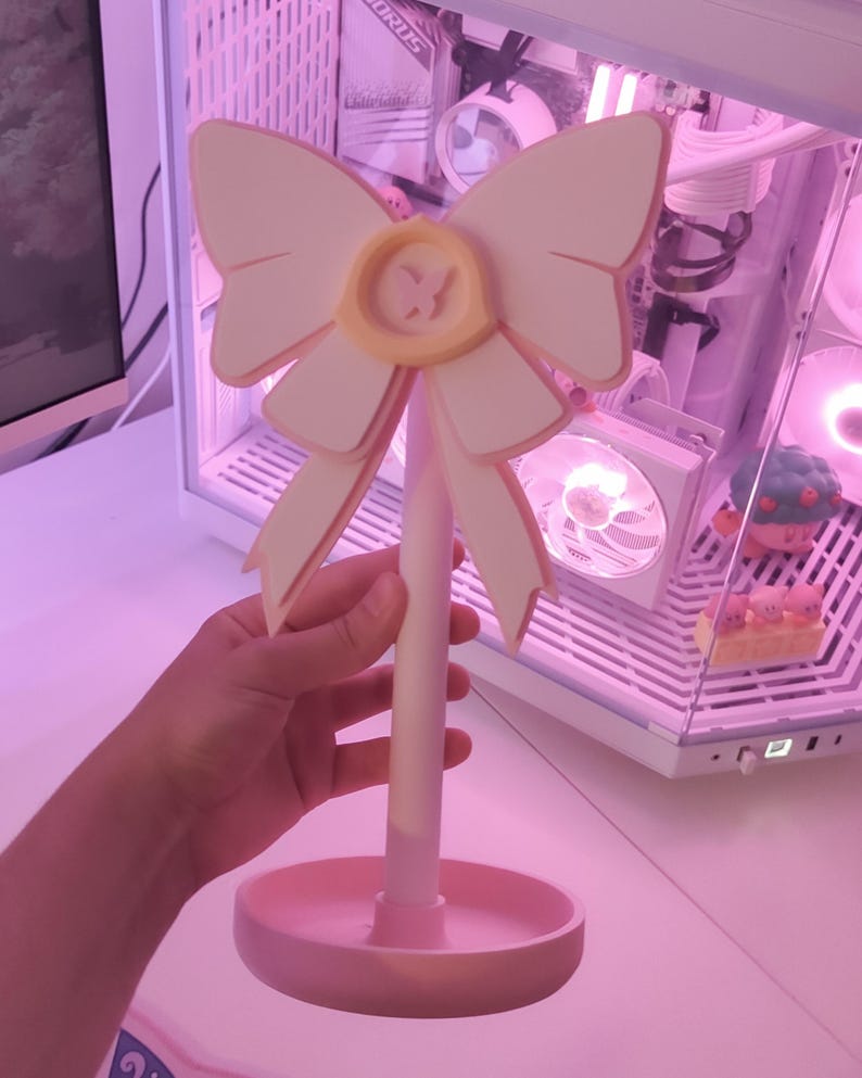 May include: A pink and white decorative stand with a large bow and a small yellow circle in the center. The stand has a round base and a tall, slender stem.