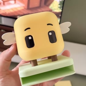 May include: A yellow, cartoon-like clip with a square head, black eyes, and small white wings. The clip has brown legs and a light green base. The design is whimsical and playful.