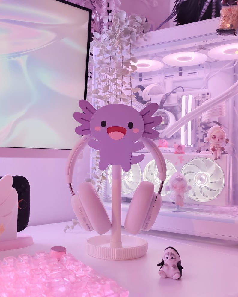 May include: A white headset rests on a decorative stand featuring a purple axolotl design. The stand is on a white surface, with a small penguin figurine and other decorative items in the background. The overall aesthetic is a soft, pastel pink.