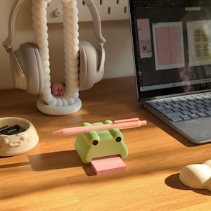May include: A green frog-shaped sticky note holder with a pink sticky note and a pink pen on top. The holder has googly eyes and a pink tongue.