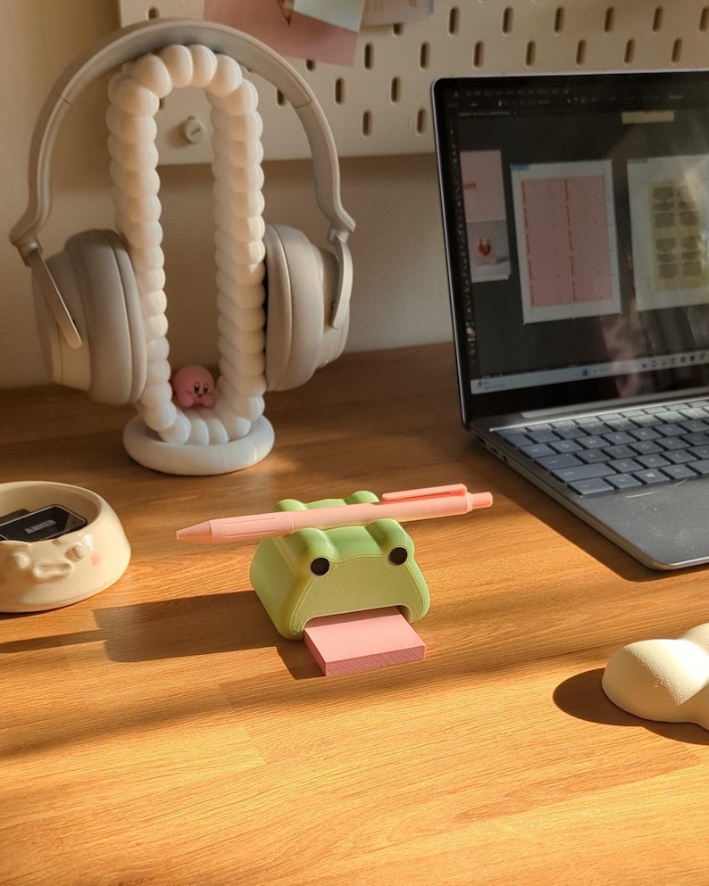 May include: A green frog-shaped sticky note holder with a pink sticky note and a pink pen on top. The holder has googly eyes and a pink tongue.
