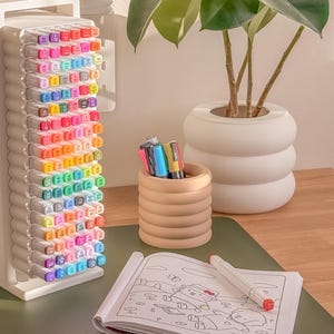May include: A white marker holder filled with colorful markers, a small wooden pen holder, a coloring book, and a white plant pot with a green plant. The markers are arranged by color, and the coloring book is open to a partially colored page.