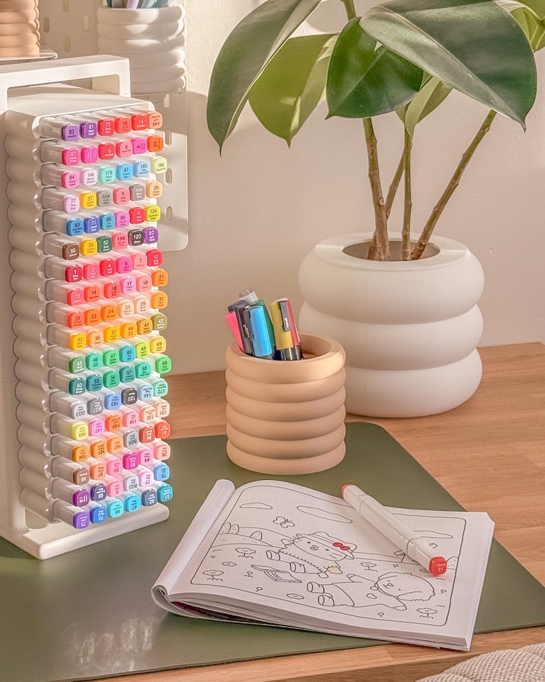May include: A white marker holder filled with colorful markers, a small wooden pen holder, a coloring book, and a white plant pot with a green plant. The markers are arranged by color, and the coloring book is open to a partially colored page.