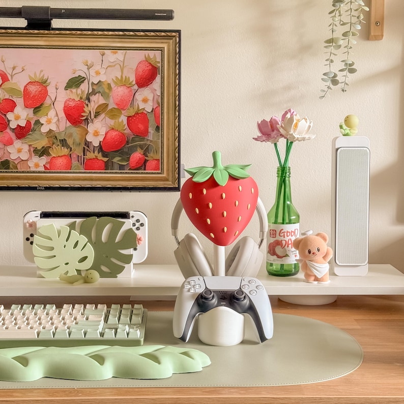 May include: A decorative display featuring a red strawberry-shaped headphone stand, a white game controller, and a Nintendo Switch. The scene includes a framed strawberry painting, a green vase with flowers, and a small teddy bear figurine.