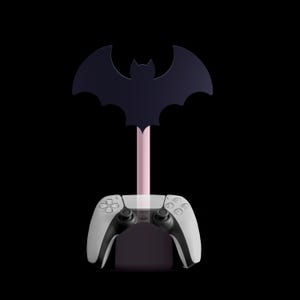K&ouml;nnte beinhalten: Ein schwarzer, fledermausf&ouml;rmiger Controller-St&auml;nder mit rosa Stiel, der einen wei&szlig;-schwarzen Gamecontroller h&auml;lt. Der St&auml;nder ist f&uuml;r einen Gamecontroller konzipiert und hat eine Fledermaus-Silhouette.
