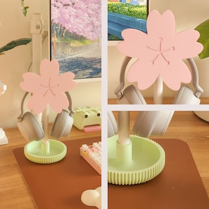 May include: A light green and pink headset stand with a cherry blossom design. The stand has two hooks for headphones and a base with a decorative pattern.