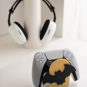 May include: A white and gray gaming controller with a black bat and yellow moon decoration. White headphones with black accents are in the background. The controller has a black center and gray buttons.