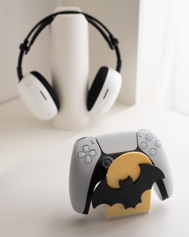 May include: A white and gray gaming controller with a black bat and yellow moon decoration. White headphones with black accents are in the background. The controller has a black center and gray buttons.