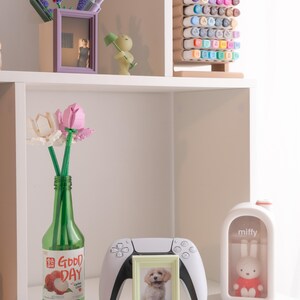 Pu&ograve; includere: Una mensola espone oggetti decorativi: una bottiglia verde con testo "Good Day", una foto di cucciolo incorniciata, un controller di gioco bianco e una statuina di coniglio "miffy". Altri oggetti includono una foto incorniciata viola, una statuina verde e un portamarker in legno.