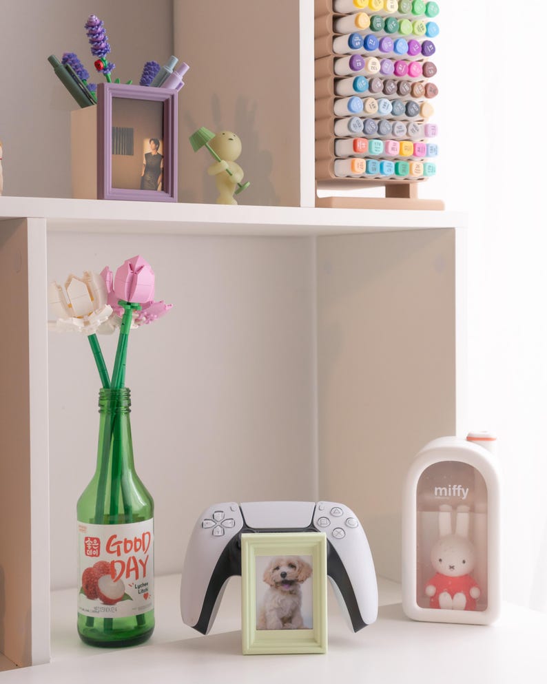 Pu&ograve; includere: Una mensola espone oggetti decorativi: una bottiglia verde con testo "Good Day", una foto di cucciolo incorniciata, un controller di gioco bianco e una statuina di coniglio "miffy". Altri oggetti includono una foto incorniciata viola, una statuina verde e un portamarker in legno.